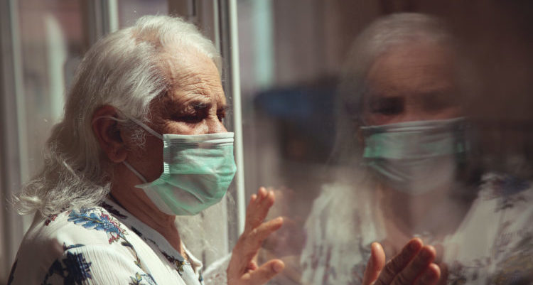 A old woman or grandma is wearing a respirator or surgical mask and looking out of the window